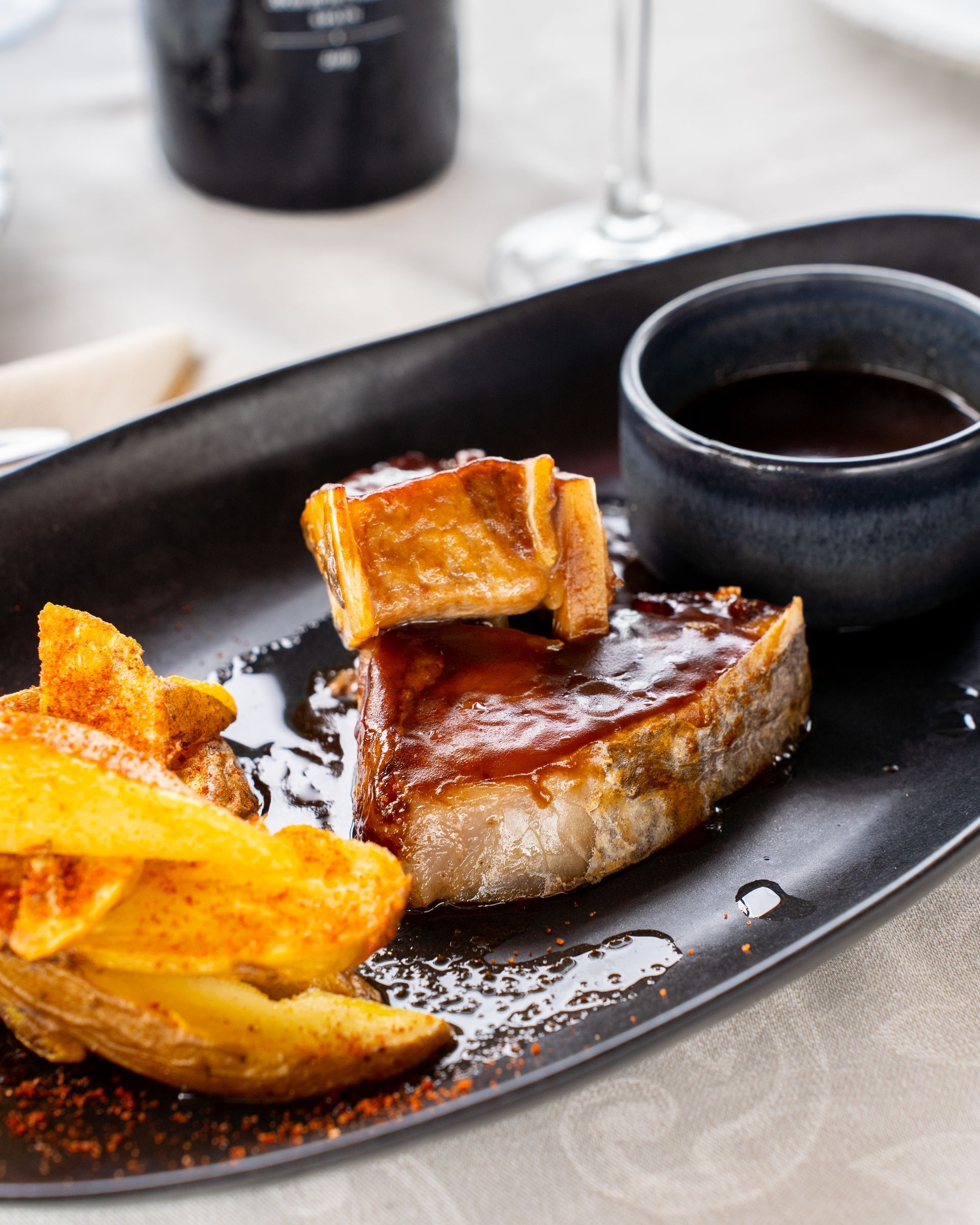 Filete, patatas crujientes y salsa en un plato negro. Junto al filete hay un plato pequeño con salsa.