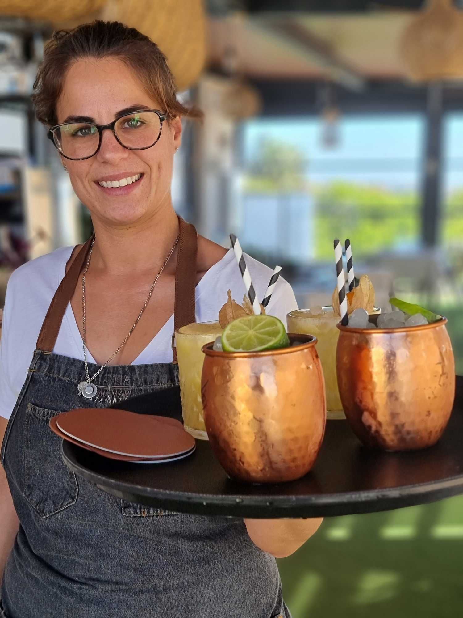 Mujer con gafas, sosteniendo una bandeja de cócteles en tazas de cobre; entorno al aire libre.