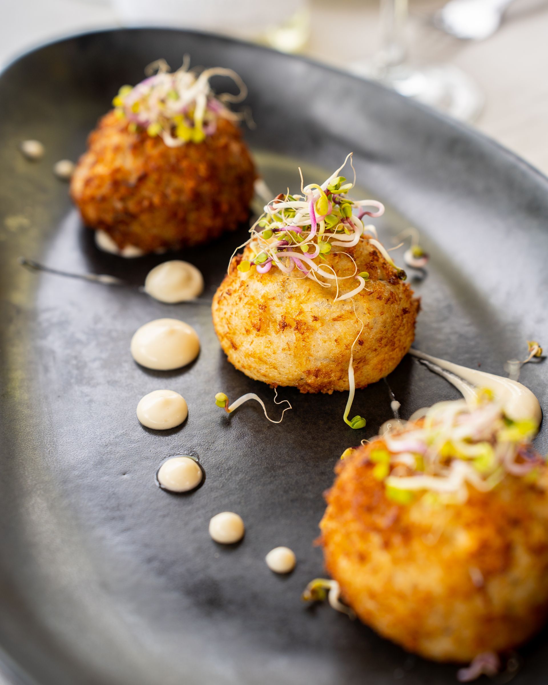 Tres croquetas de patata fritas doradas sobre un plato negro, adornadas con brotes y puntos de salsa.