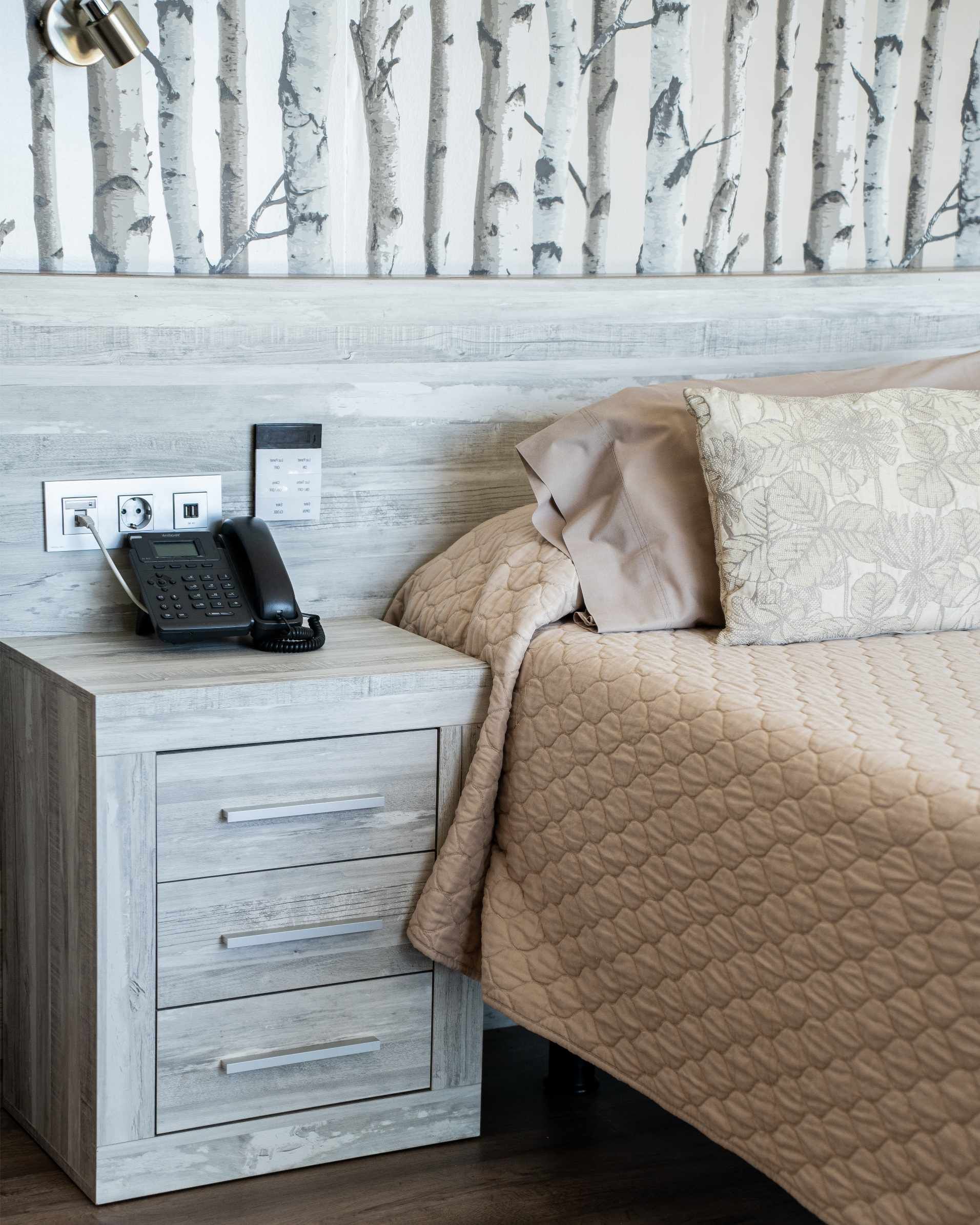 Mesilla de noche con teléfono, cama con edredón beige, papel pintado color abedul.