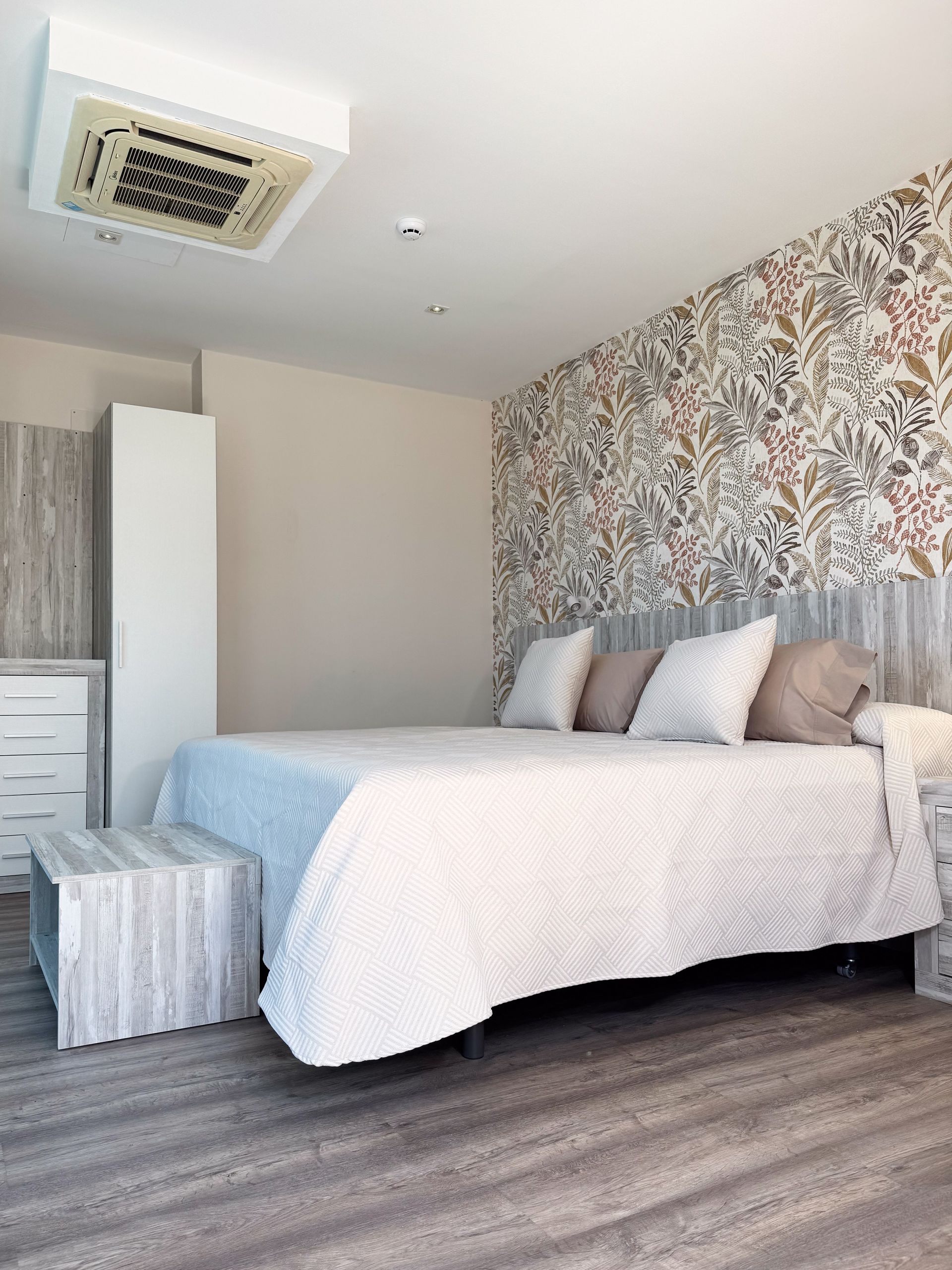 Dormitorio con papel pintado estampado, cama con funda blanca, almohadas y banco de madera.