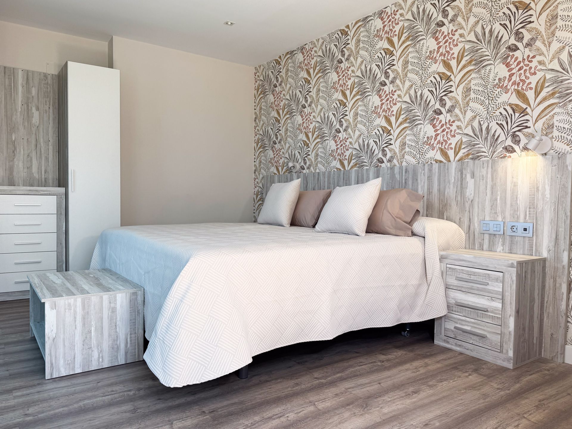 Dormitorio con cama doble, papel pintado estampado, tonos neutros, detalles en madera y pequeña mesita de noche.