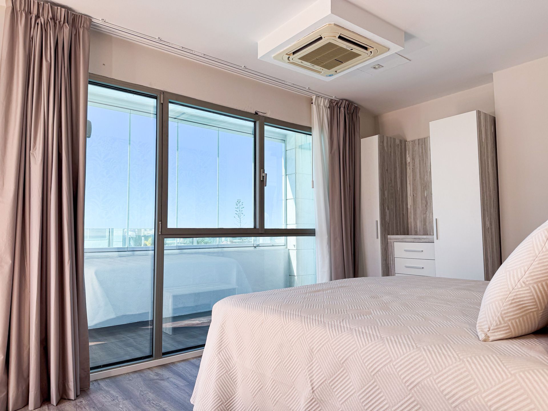 Dormitorio con gran ventanal y cortinas, cama, armarios blancos y vista al exterior.