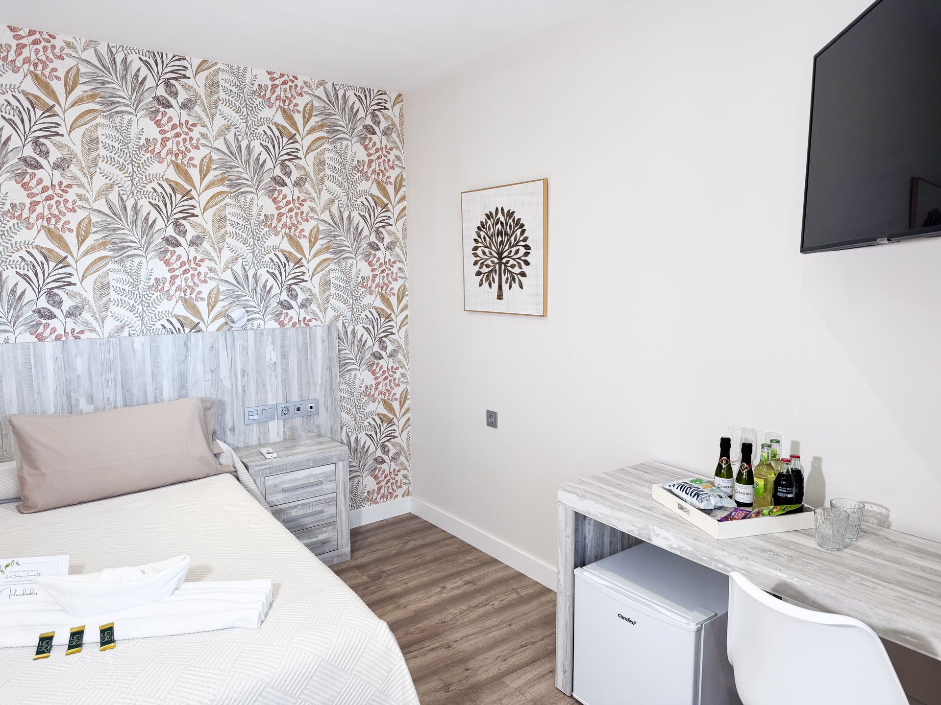 Habitación de hotel con papel tapiz floral, cama, escritorio, refrigerador pequeño y televisor montado en la pared.