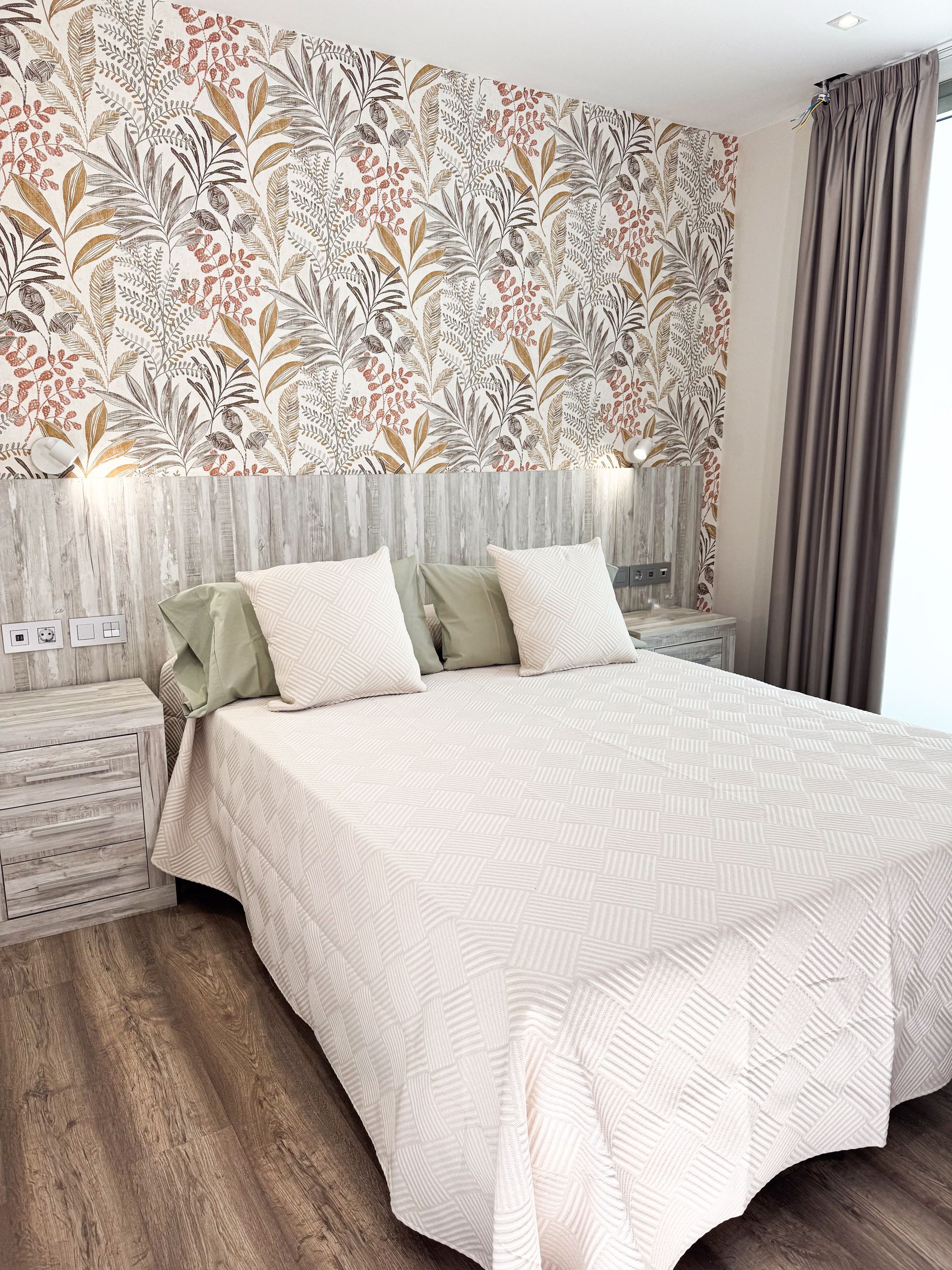 Dormitorio con papel pintado floral, cabecero de madera, colcha color crema y suelo de madera.