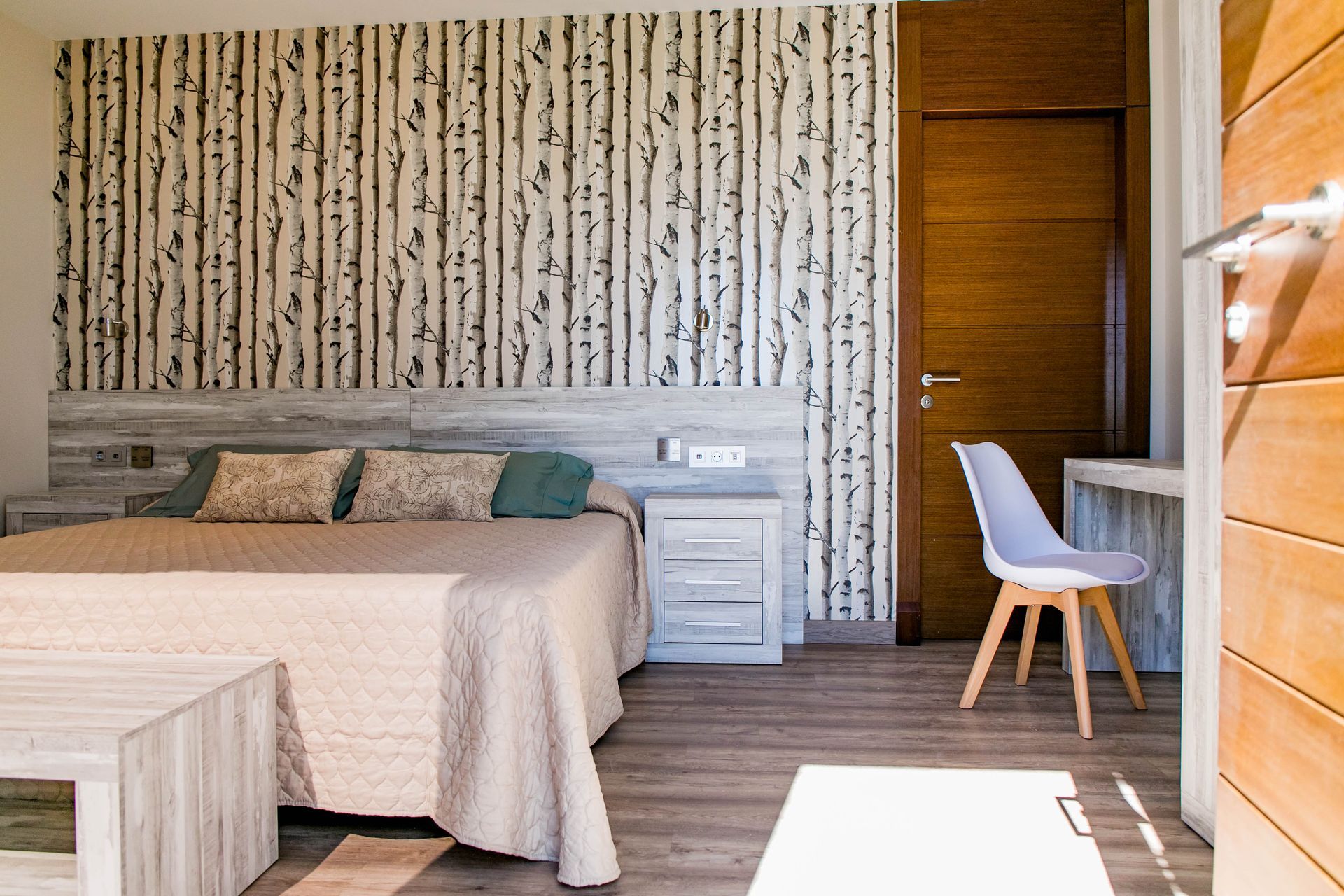 Dormitorio con papel pintado de abedul, cama, escritorio y puerta de madera clara.