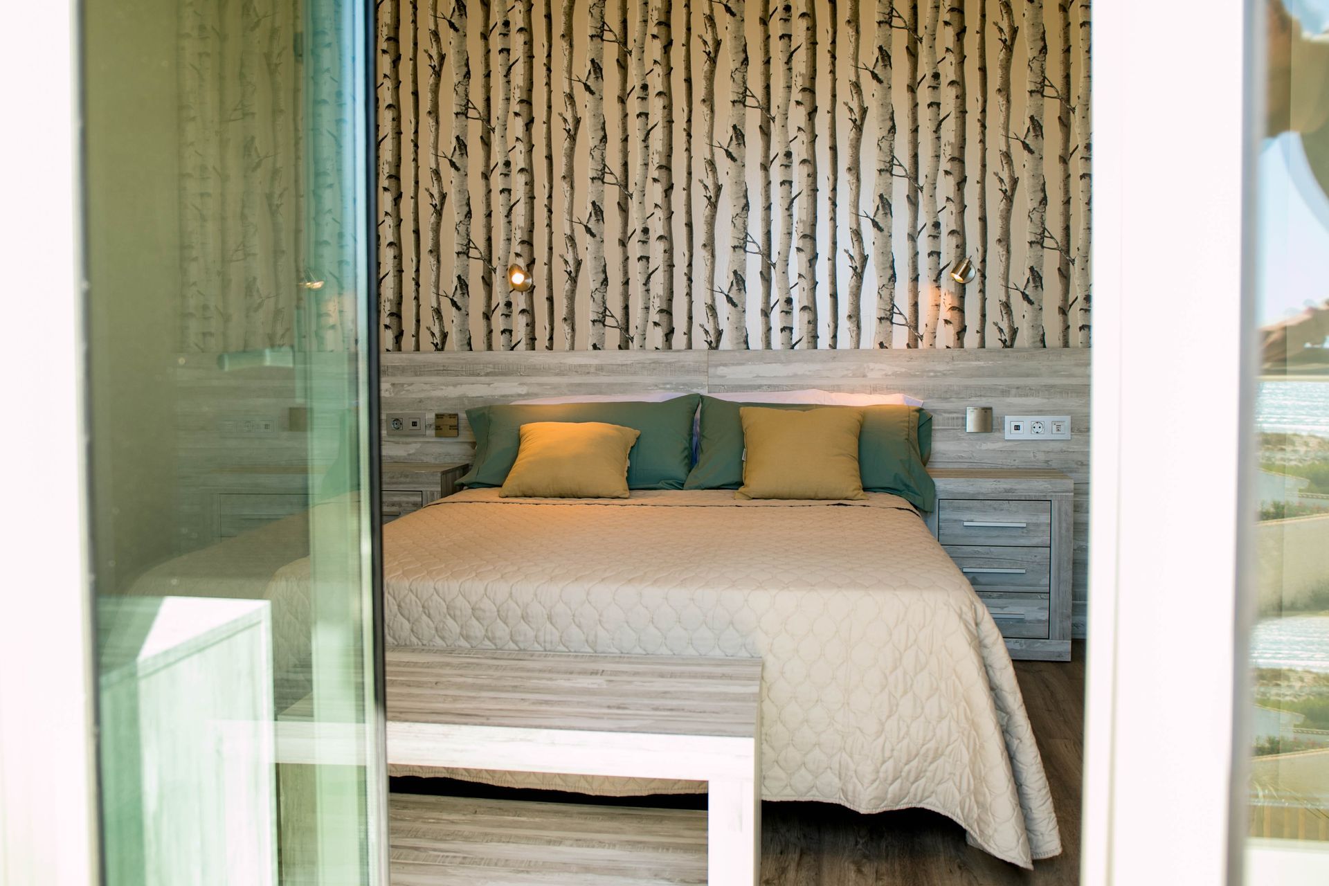 Dormitorio con cama, papel pintado con estampado de abedul, almohadas verdes y amarillas y muebles de colores claros.