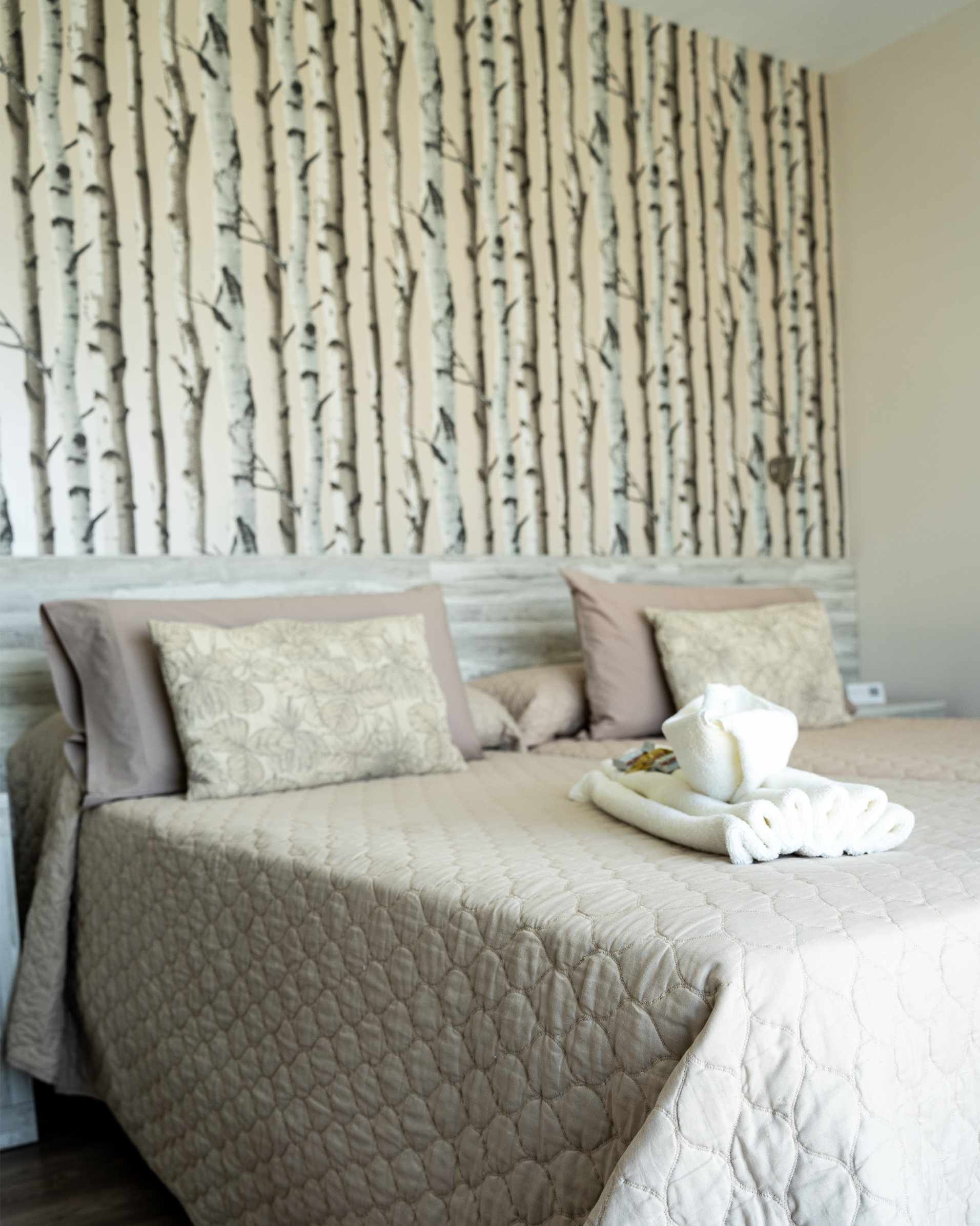 Dos camas con fundas acolchadas de color beige, almohadas estampadas y papel tapiz de abedul.