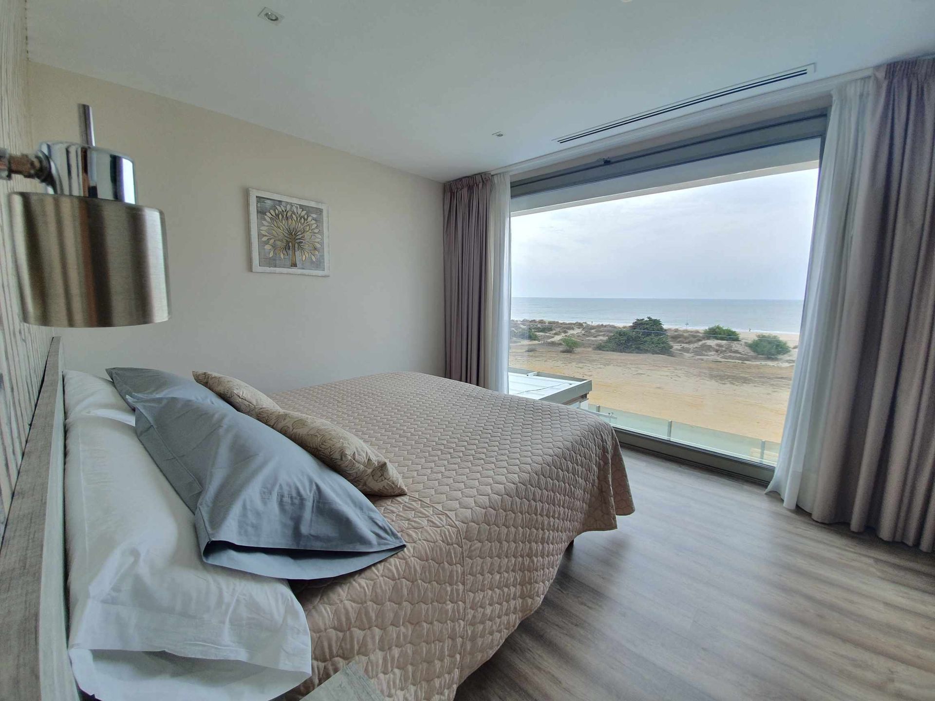 Dormitorio con cama, almohadas y gran ventanal con vistas a la playa.