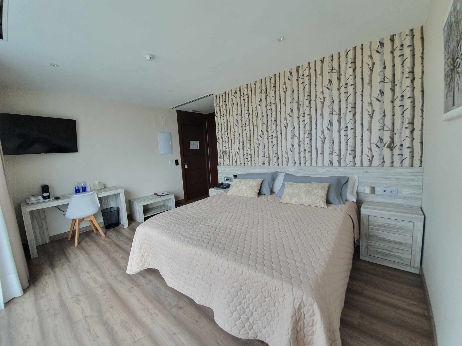 Dormitorio con pared con acento de abedul, cama con funda beige, escritorio y TV.