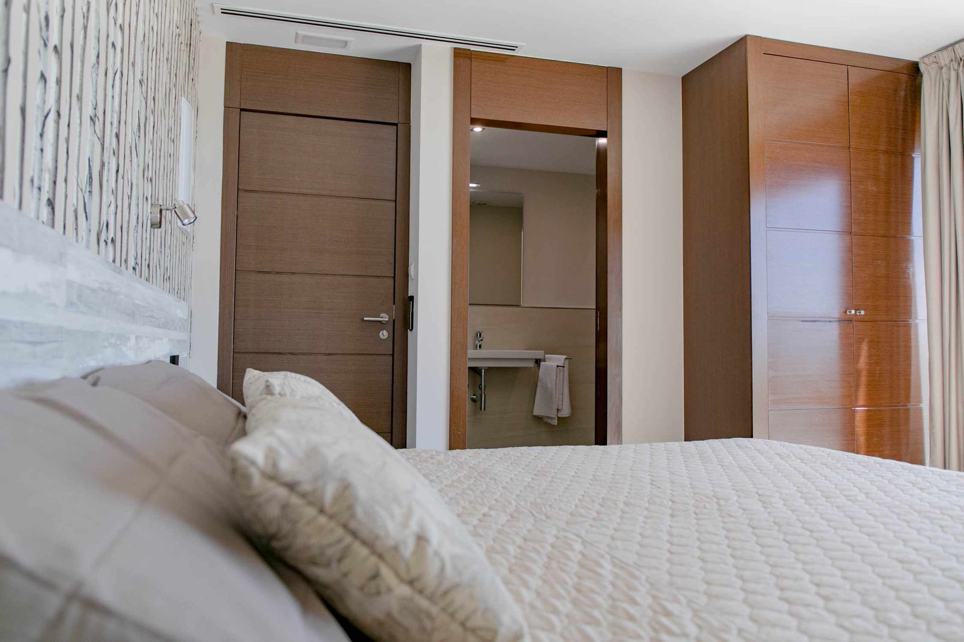 Dormitorio con cama, puertas de madera y puerta abierta a un baño con lavabo.