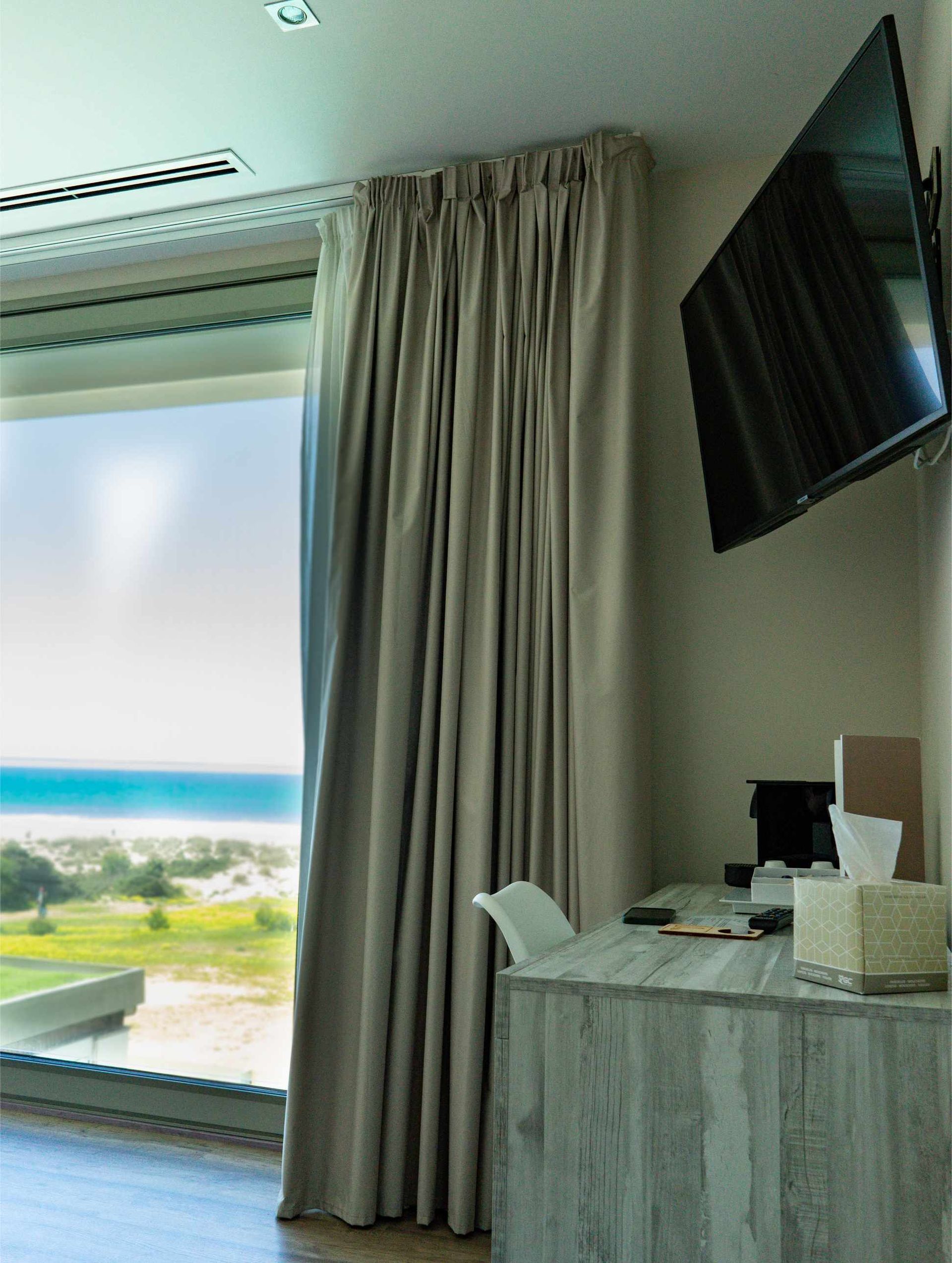 Una habitación de hotel con vistas. Una gran ventana, cortinas grises, un escritorio y un televisor.