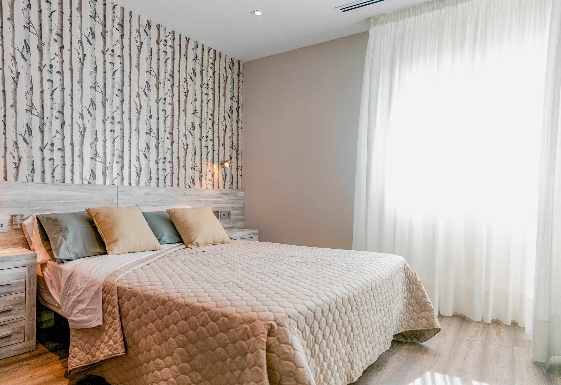 Dormitorio con papel tapiz de abedul, cama con edredón beige y cortinas blancas transparentes.