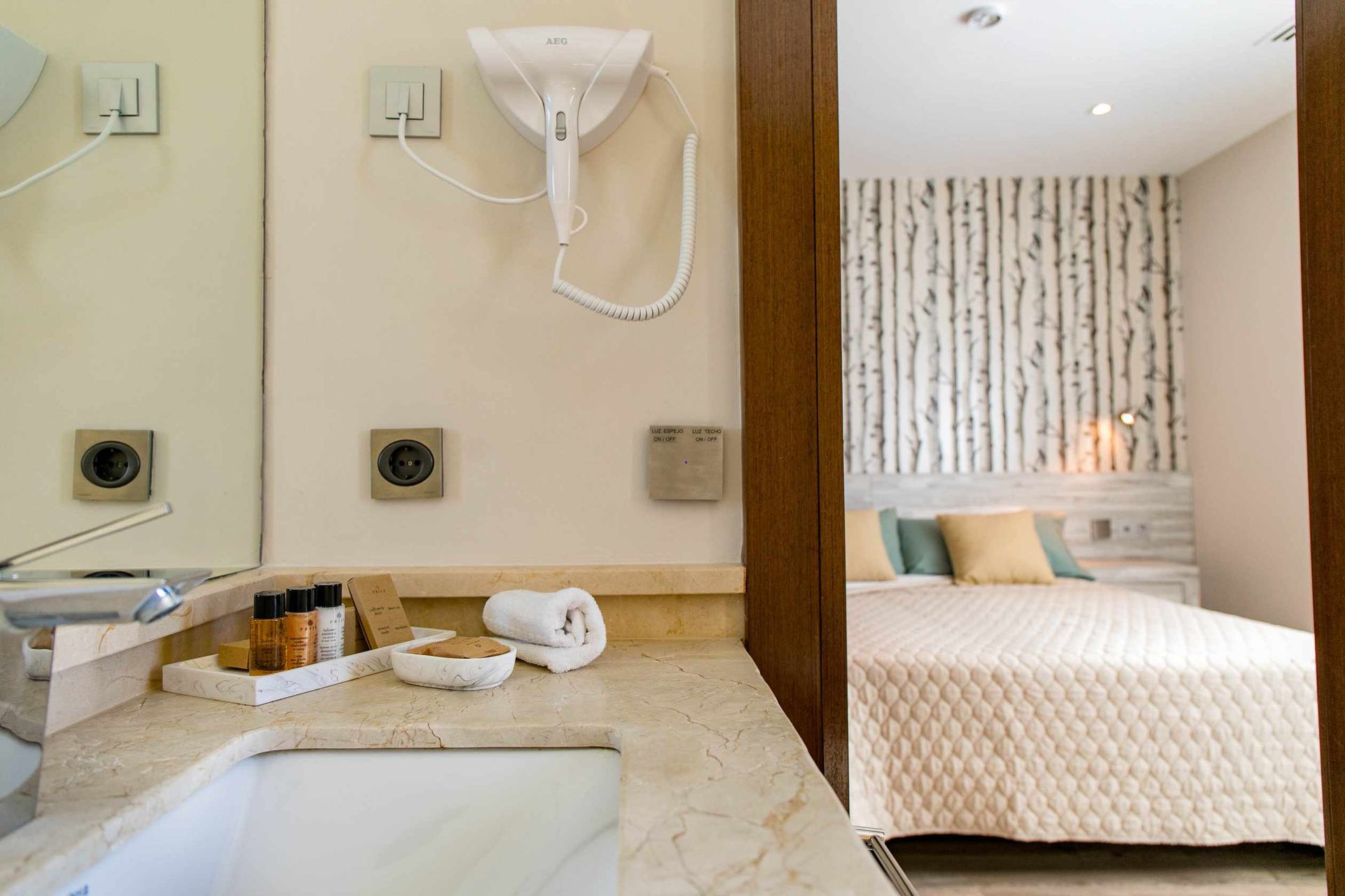 Mueble de baño con artículos de aseo, secador de pelo y vista al dormitorio con cama.