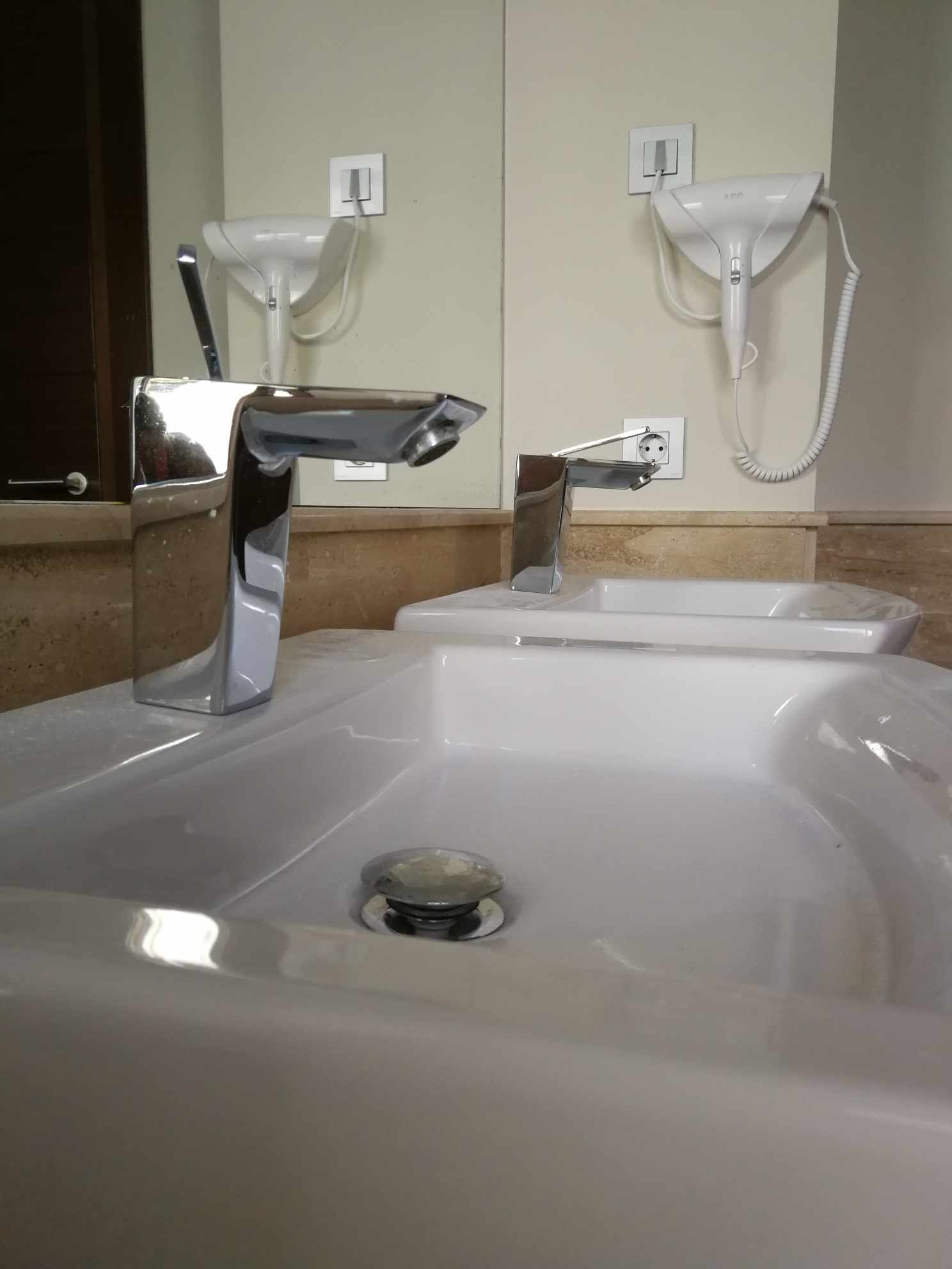 Grifo cromado sobre un lavabo blanco con reflejo de espejo; secador de pelo en la pared.