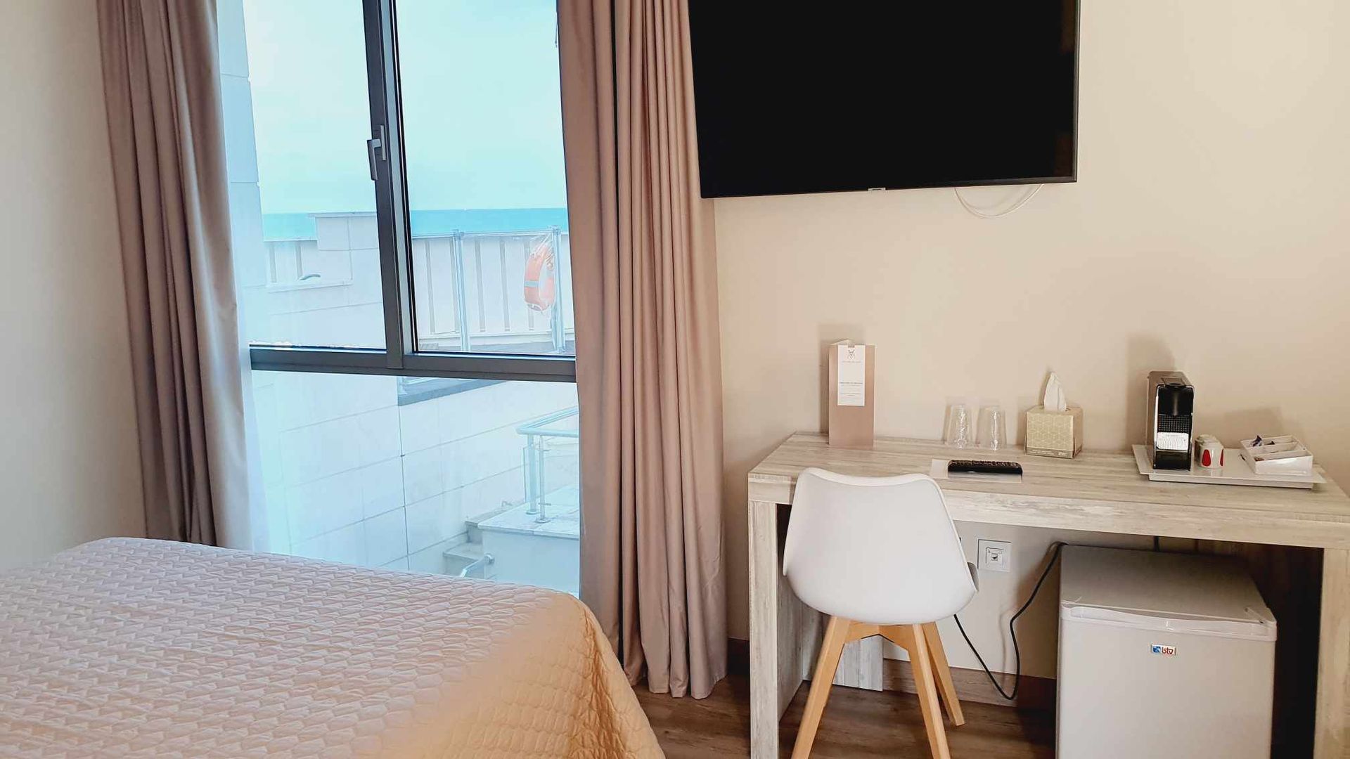 Dormitorio con cama, escritorio, ventana con vista al mar y televisor. Tonos blanco y beige.