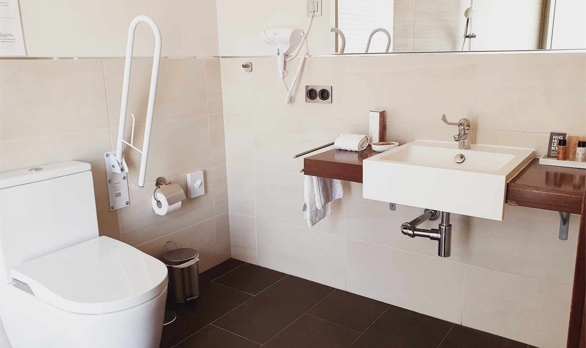 Baño accesible: Inodoro, lavabo y barra de apoyo. Paredes beige, suelo oscuro.