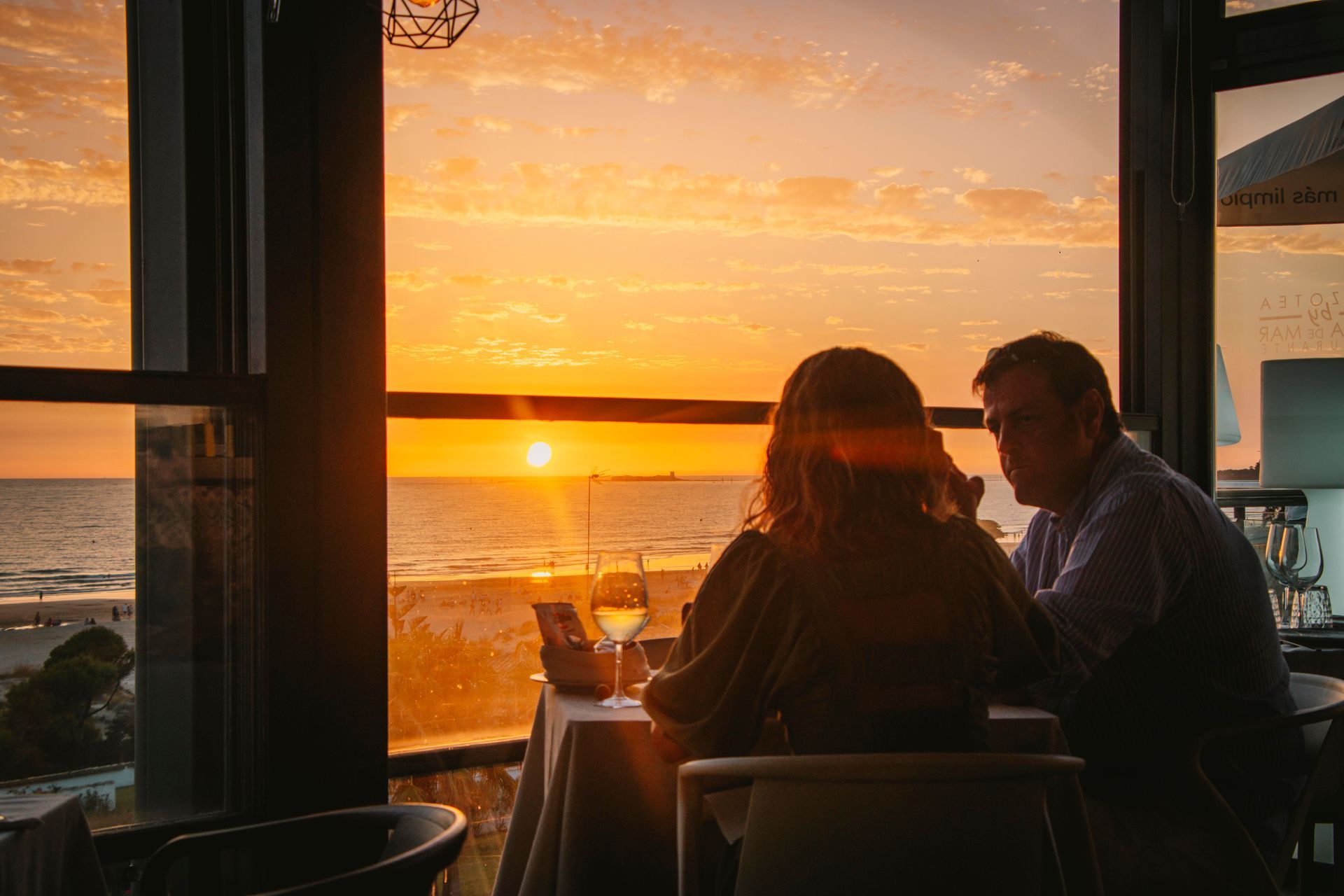 Pareja cenando, mirando la puesta de sol sobre el océano a través de la ventana; luz romántica y dorada.
