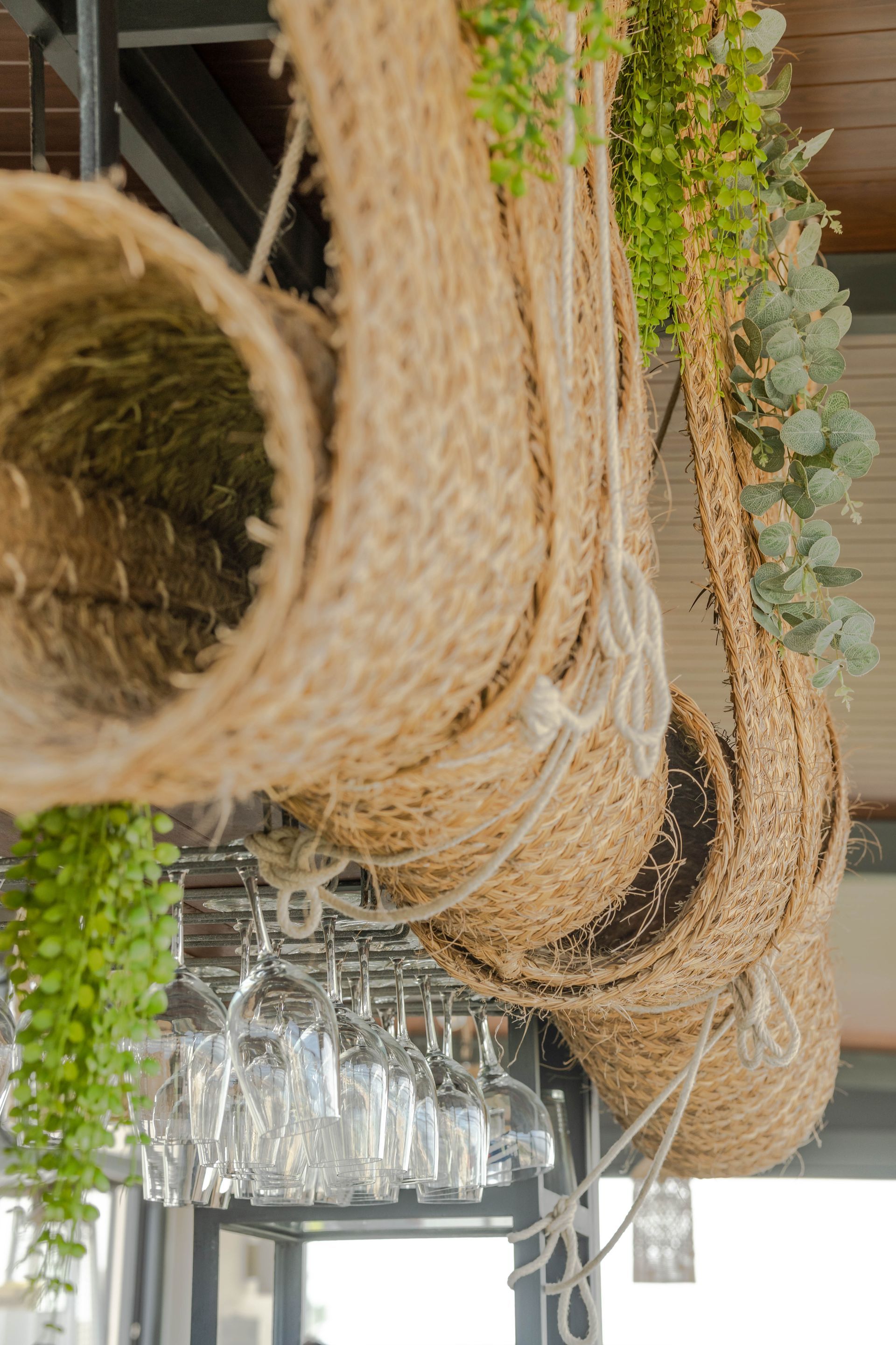 Cestas colgantes tejidas y vegetación, con copas de vino exhibidas debajo.