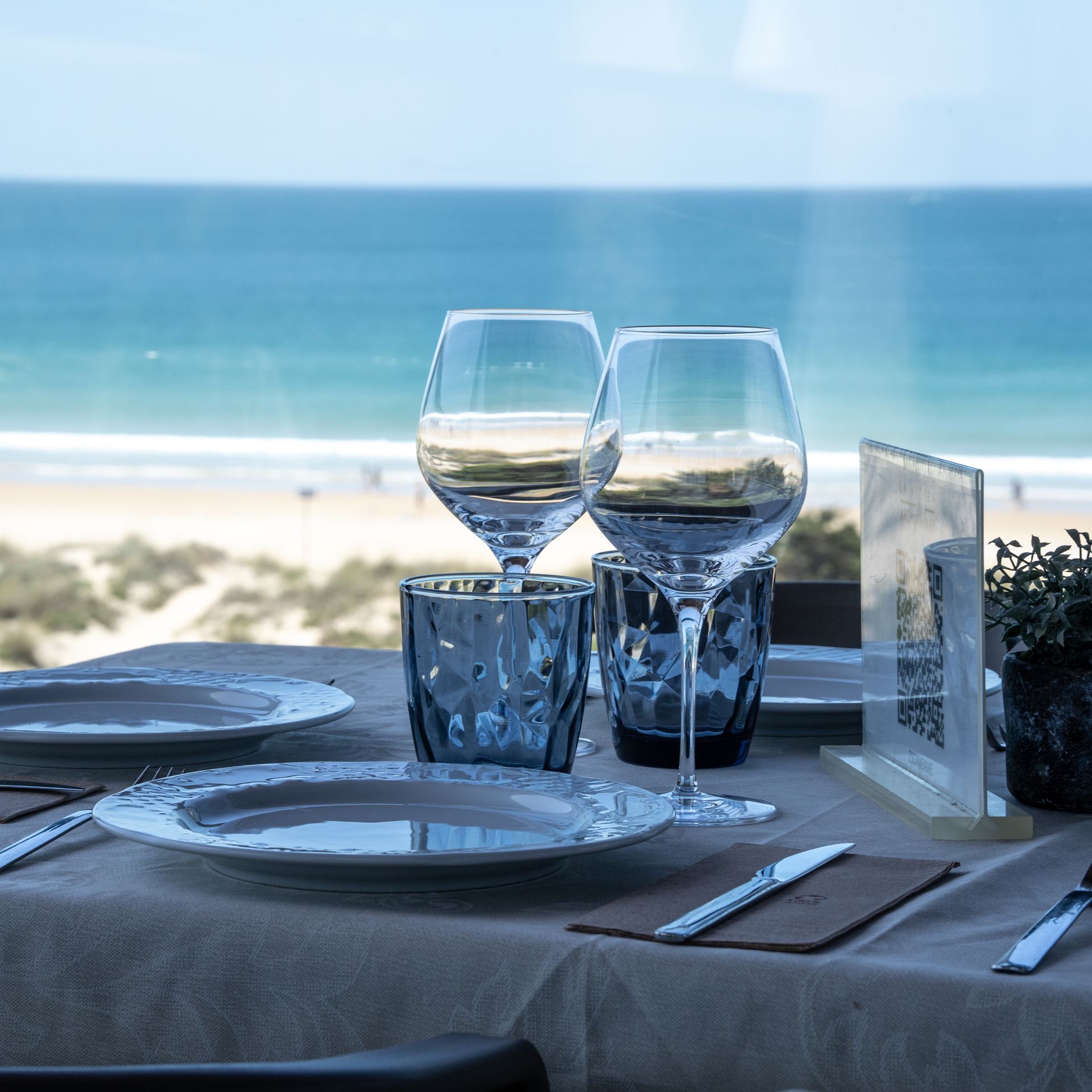 Mesa puesta para dos con vista al mar: platos, vasos y cubiertos.