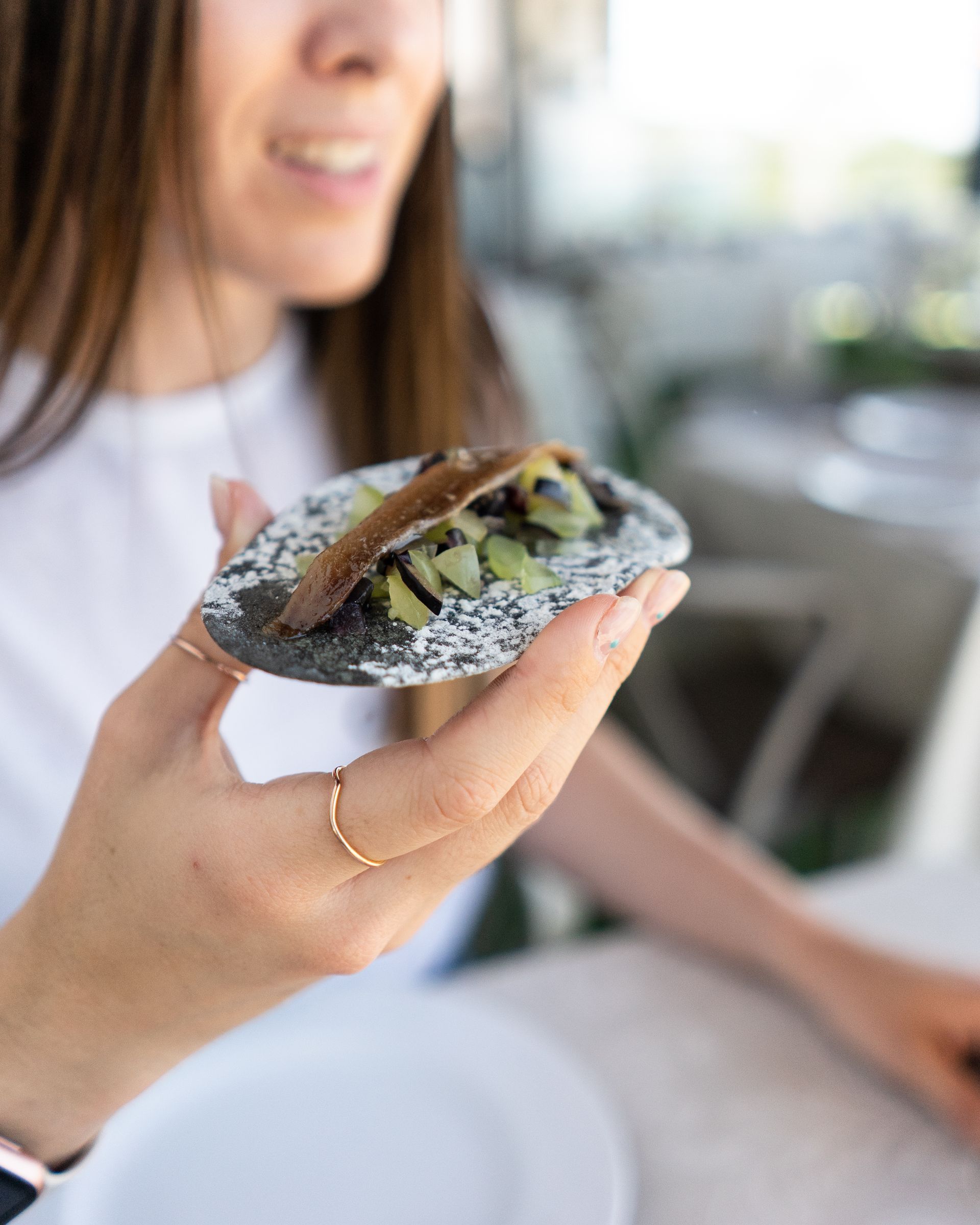 Mujer sostiene un trozo de comida con pan oscuro y aderezos; sonriendo al aire libre.