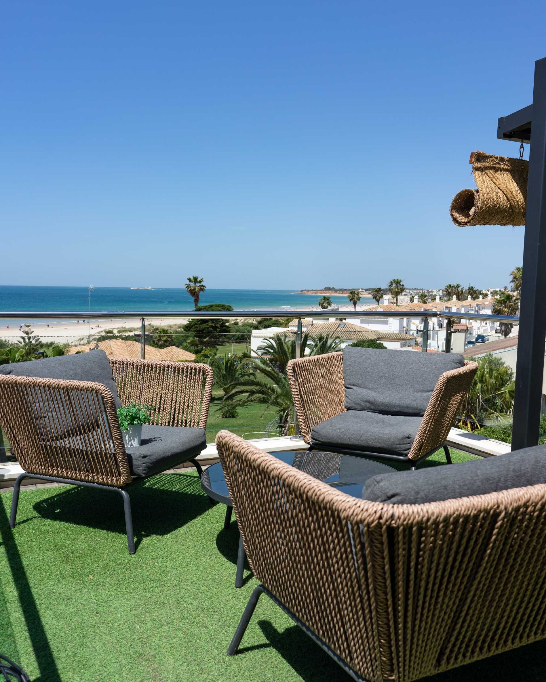 Zona de estar en la azotea con vista al mar: sillas de mimbre, césped artificial, cielo azul y playa de fondo.