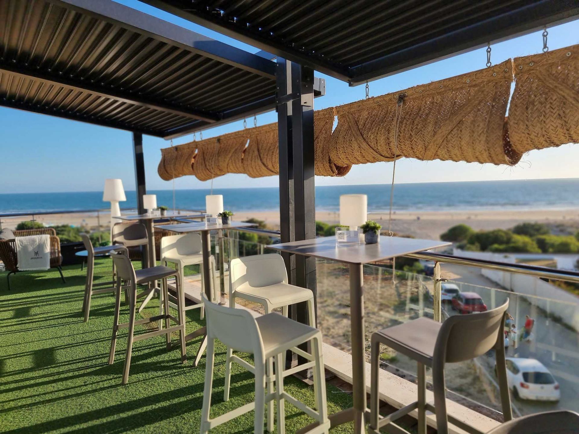 Bar en la azotea con vistas a la playa. Mesas y sillas bajo un toldo, con vistas al mar y al cielo.