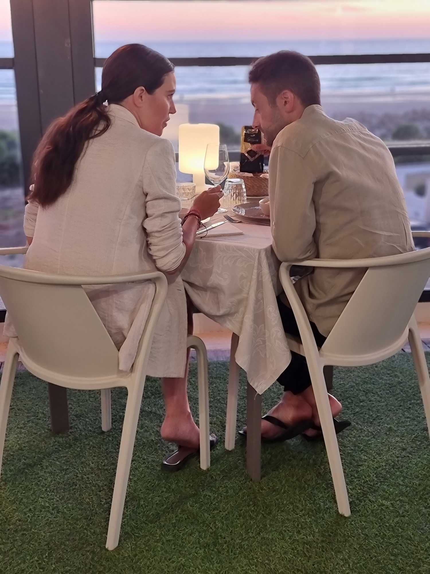Una pareja cena en un restaurante con vistas al océano al atardecer.