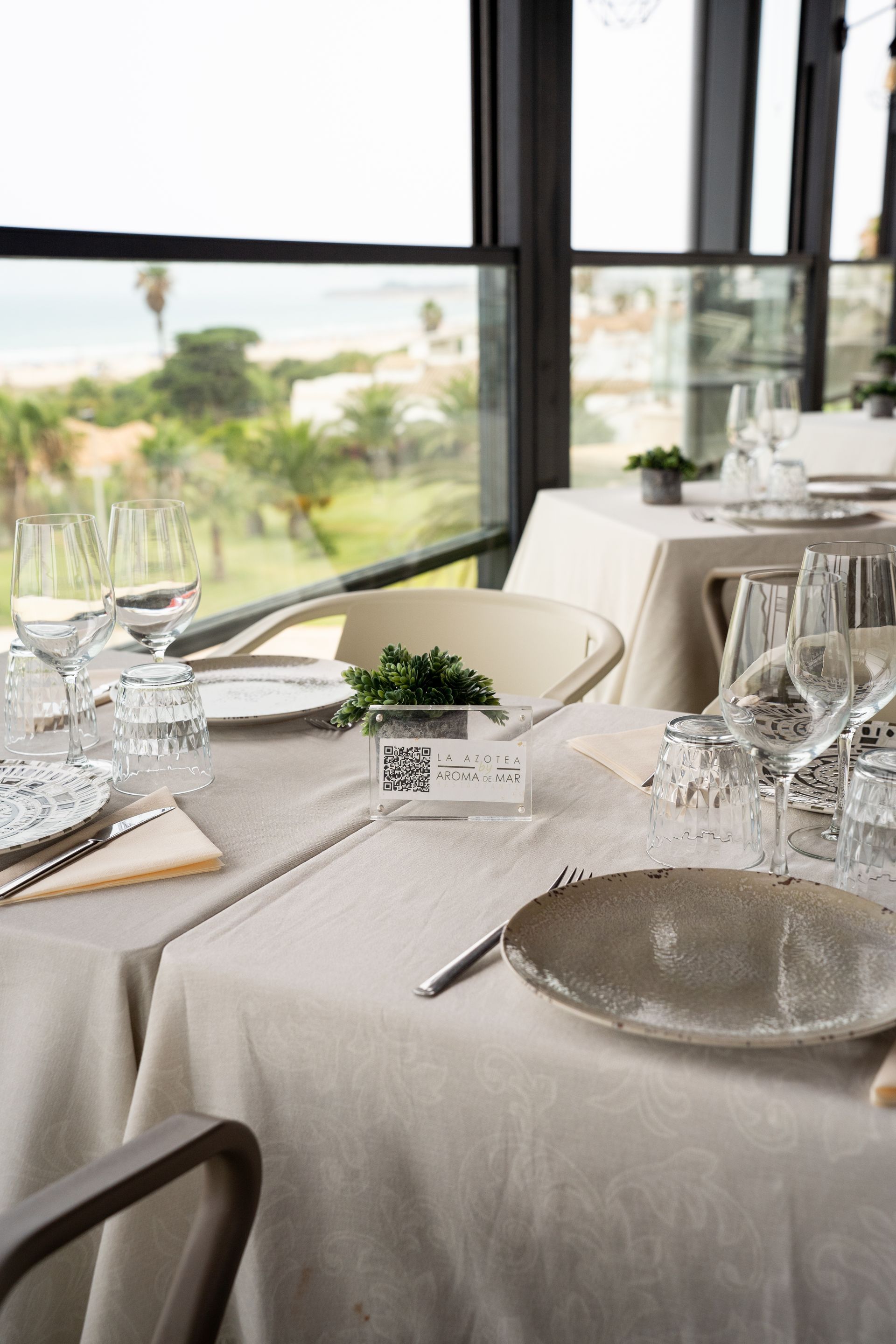 Mesa de restaurante preparada para cenar con vista al mar a través de ventana.
