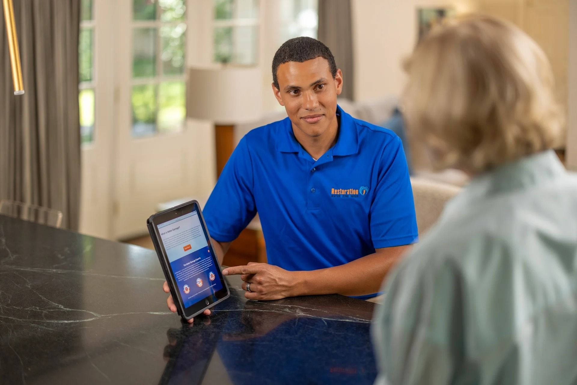 Man in blue shirt shows tablet to woman; kitchen setting.