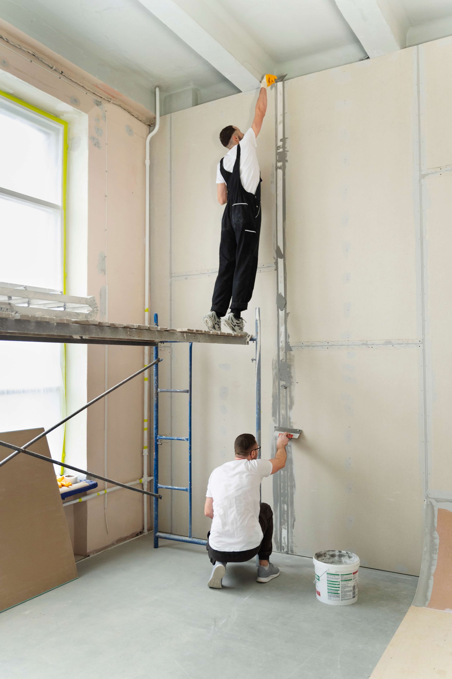 Two workers apply joint compound to drywall in a room under construction. One is on a scaffold.