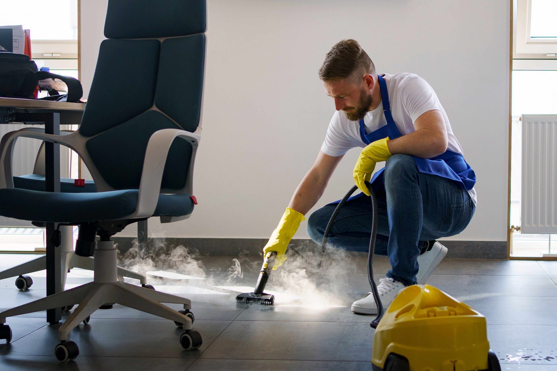 Man in apron and gloves steam-cleaning office floor near desk.