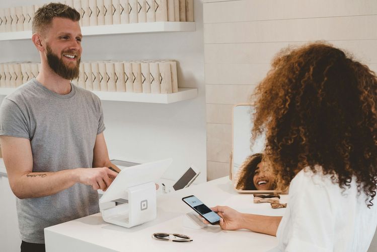 Man with beard smiles at woman paying with phone at a white counter. Store interior.