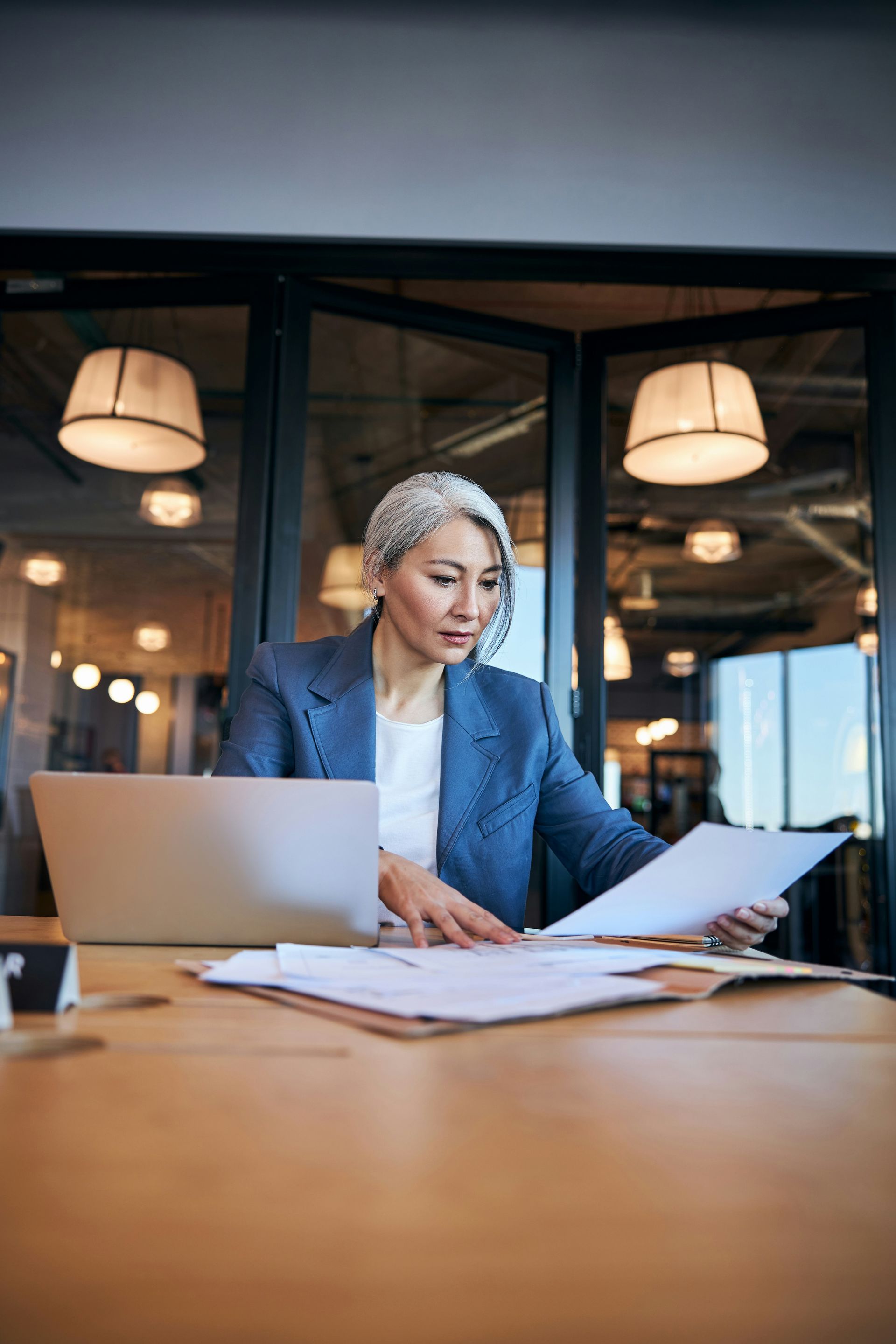 Businesswoman reviews documents at a desk, laptop nearby. She wears a blue blazer in a well-lit office.