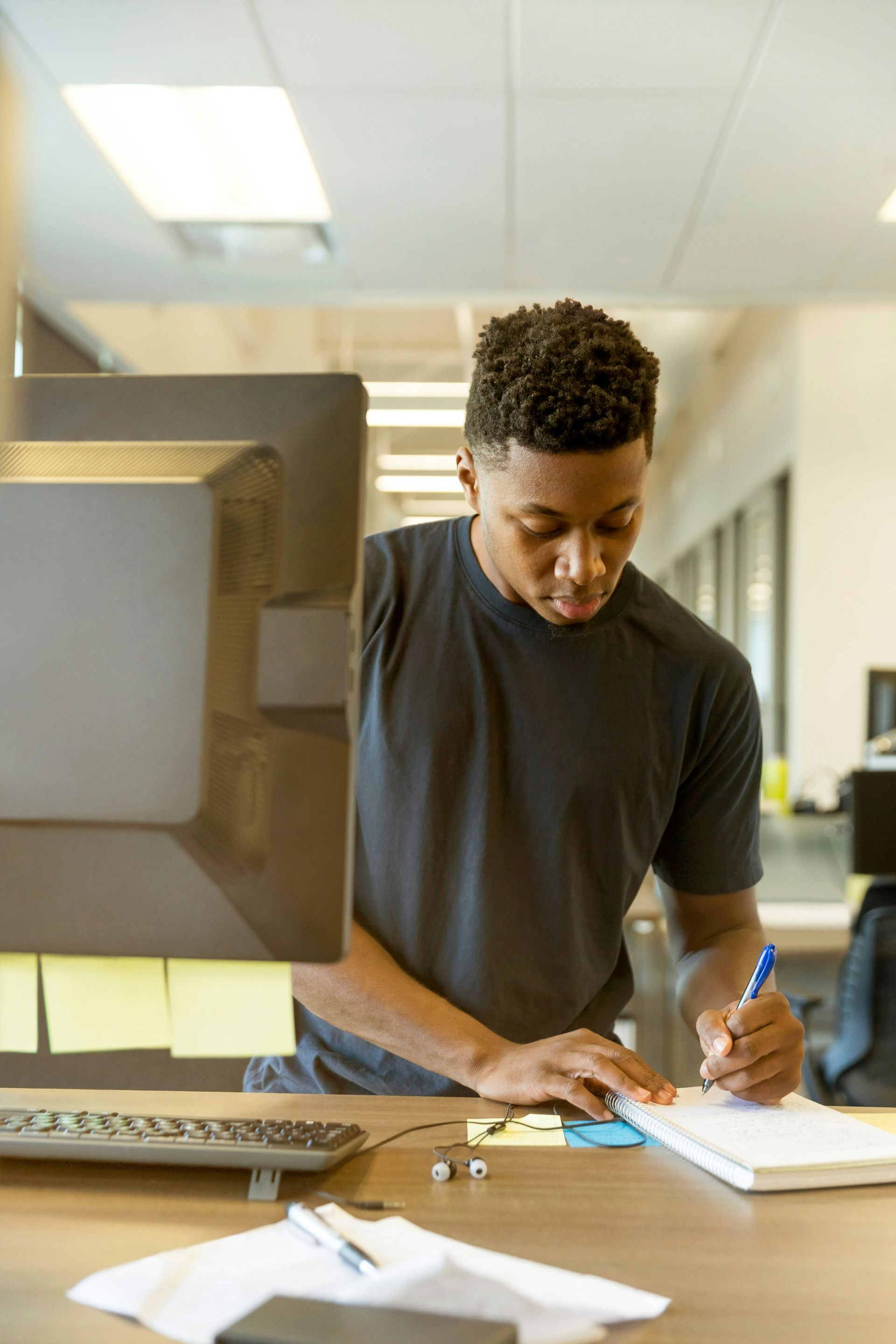 Man in dark shirt writing in notebook at a desk in an office.