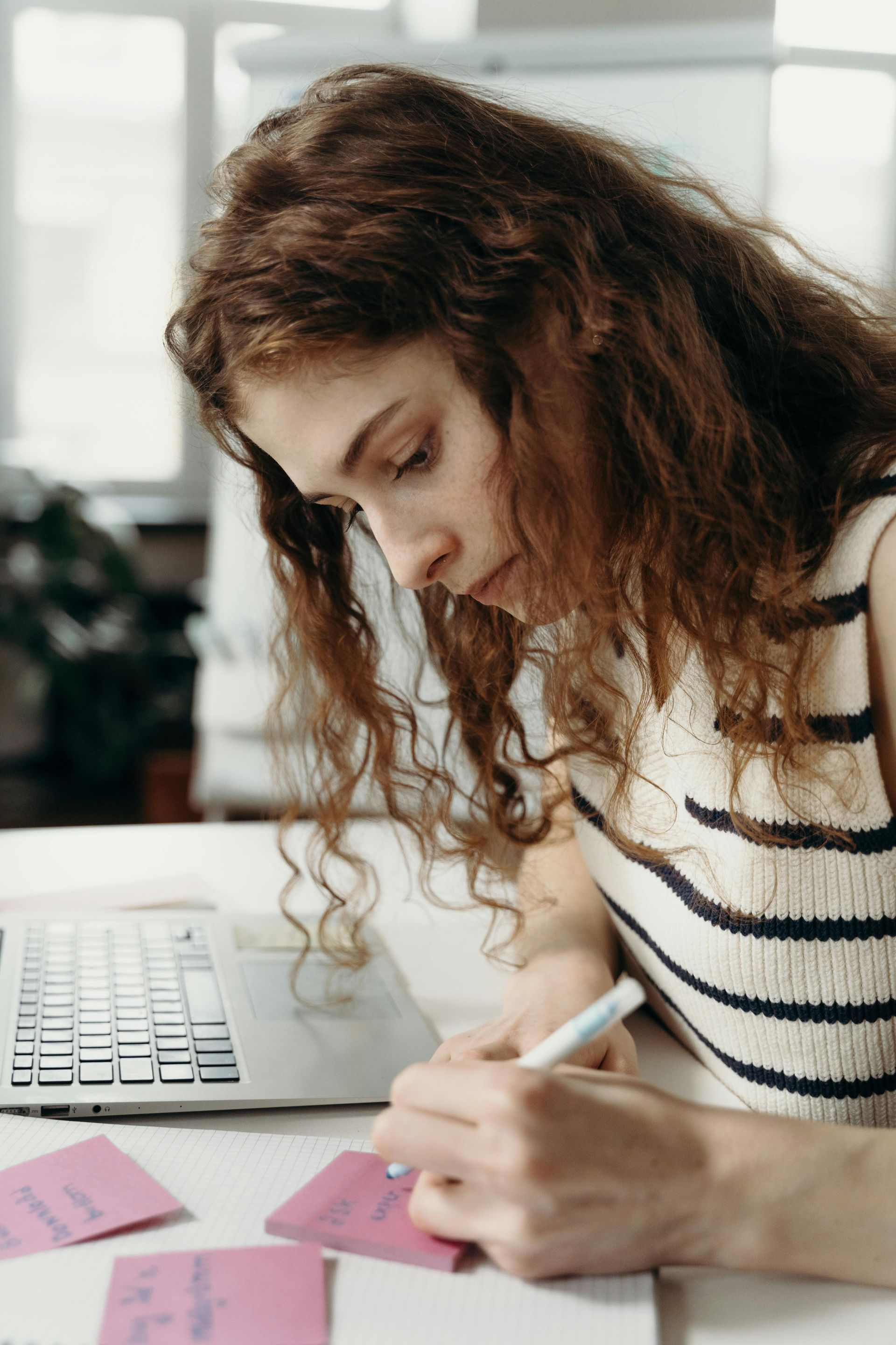 Woman with curly red hair writing on pink sticky notes at a desk with a laptop.