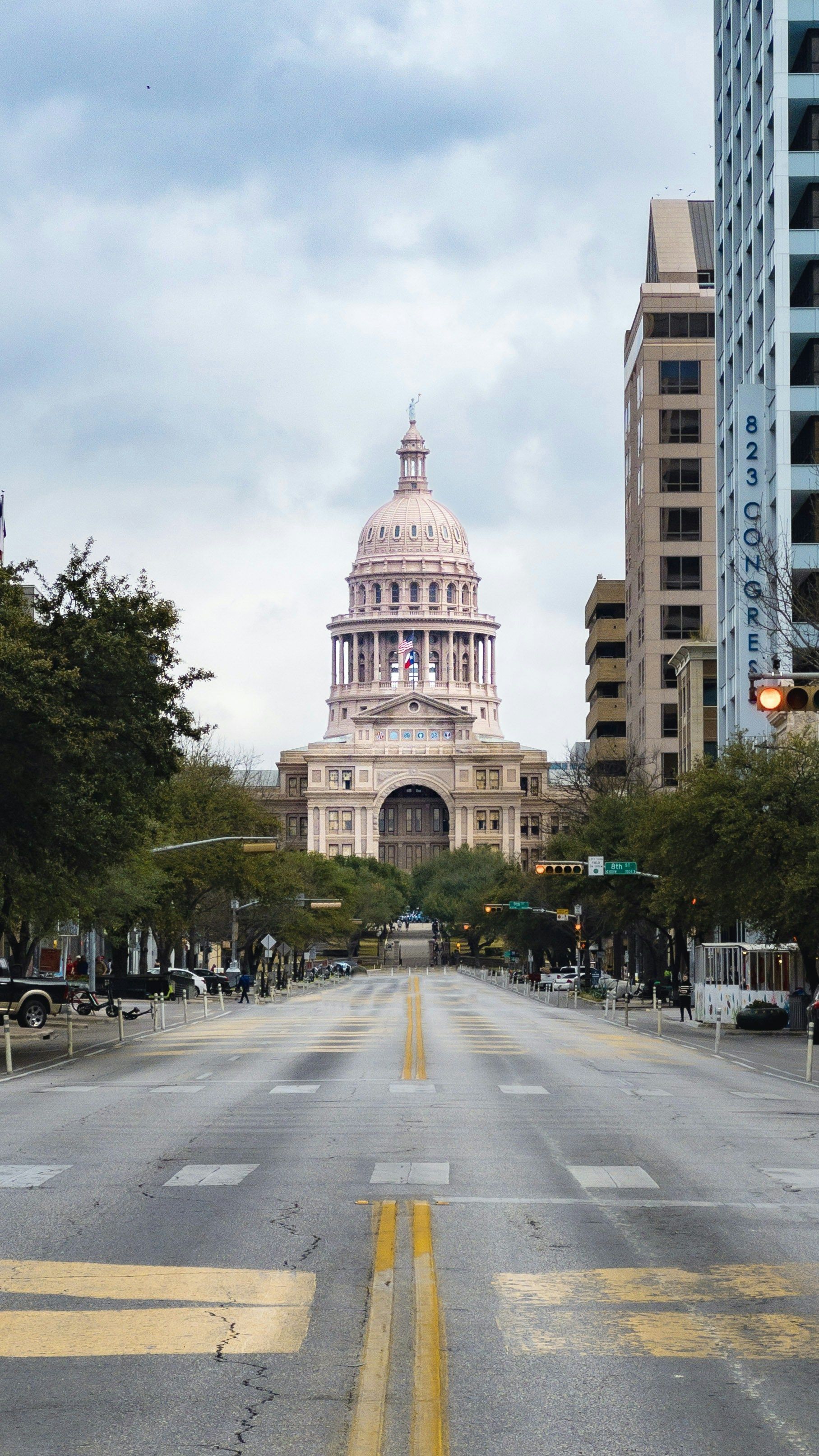 Texas State Capitol building at the end of a city street with trees on either side. Cloudy sky overhead.
