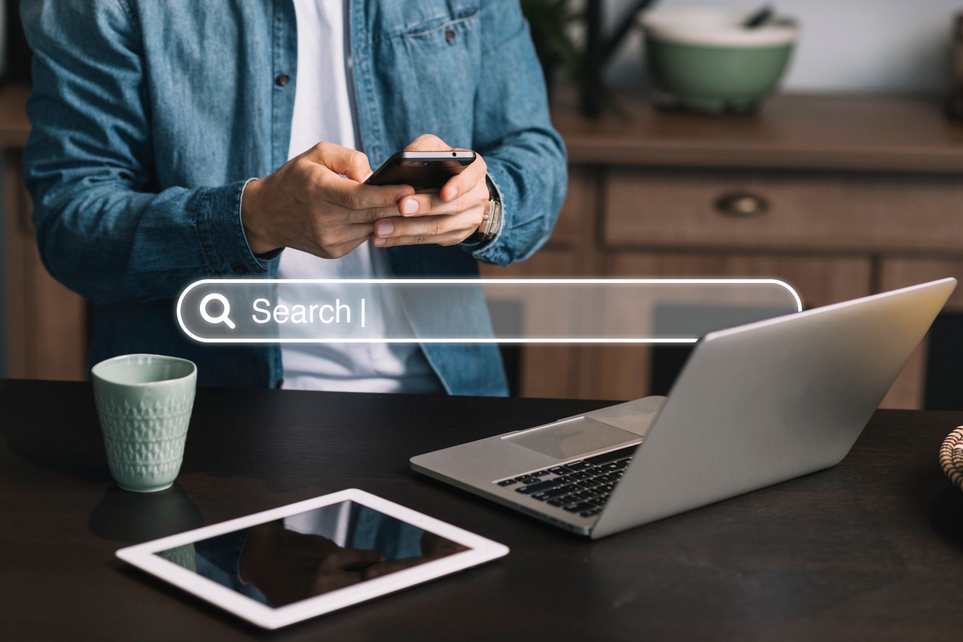 Man using a phone, with a laptop and tablet on a table, search bar overlay.