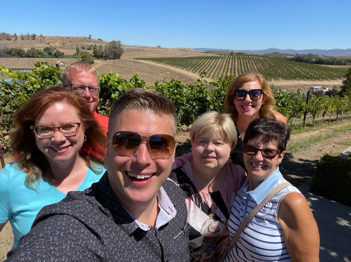 A group of people are posing for a selfie in front of a vineyard.