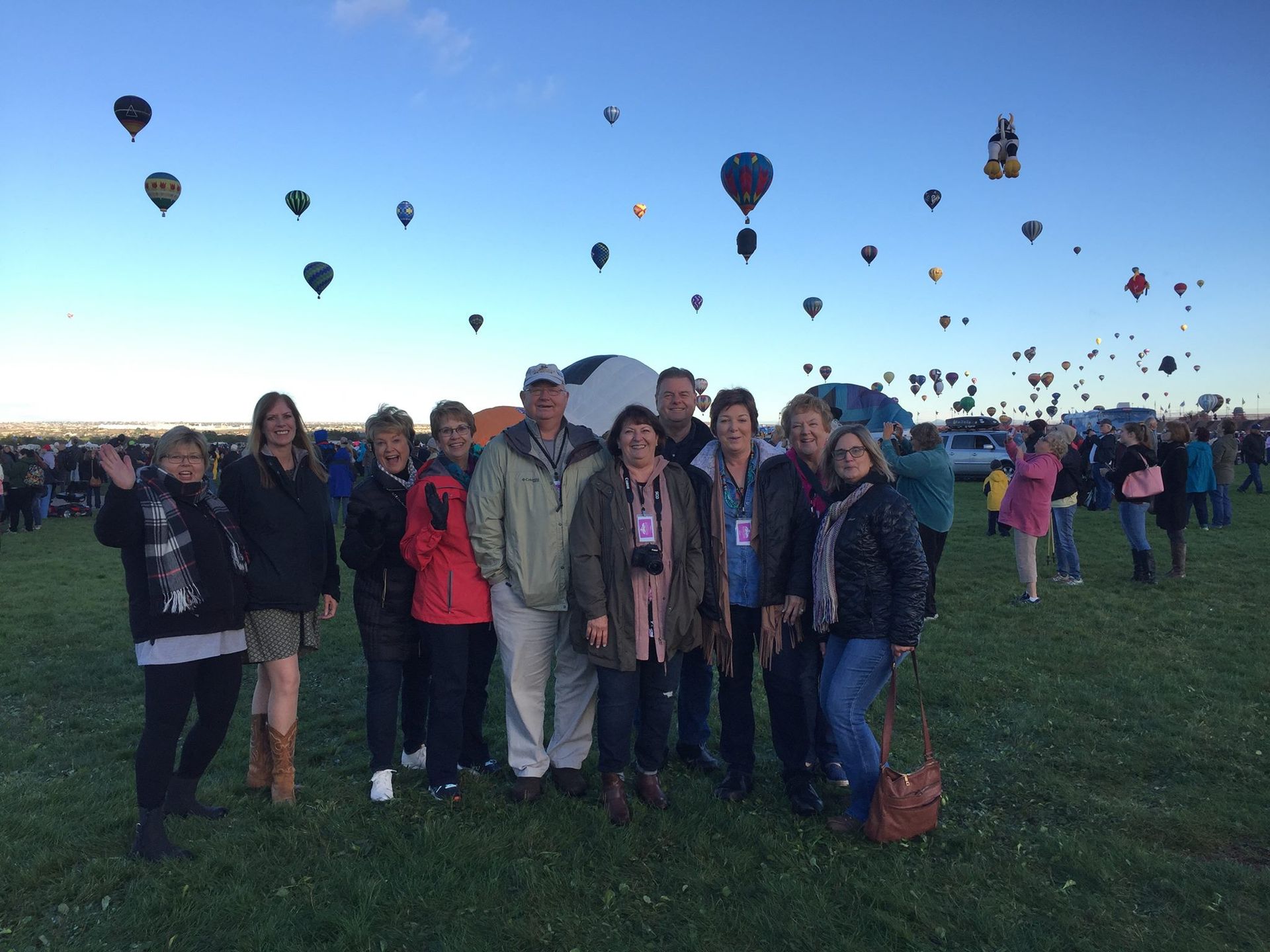 A group of people are standing in a field with hot air balloons flying in the background.