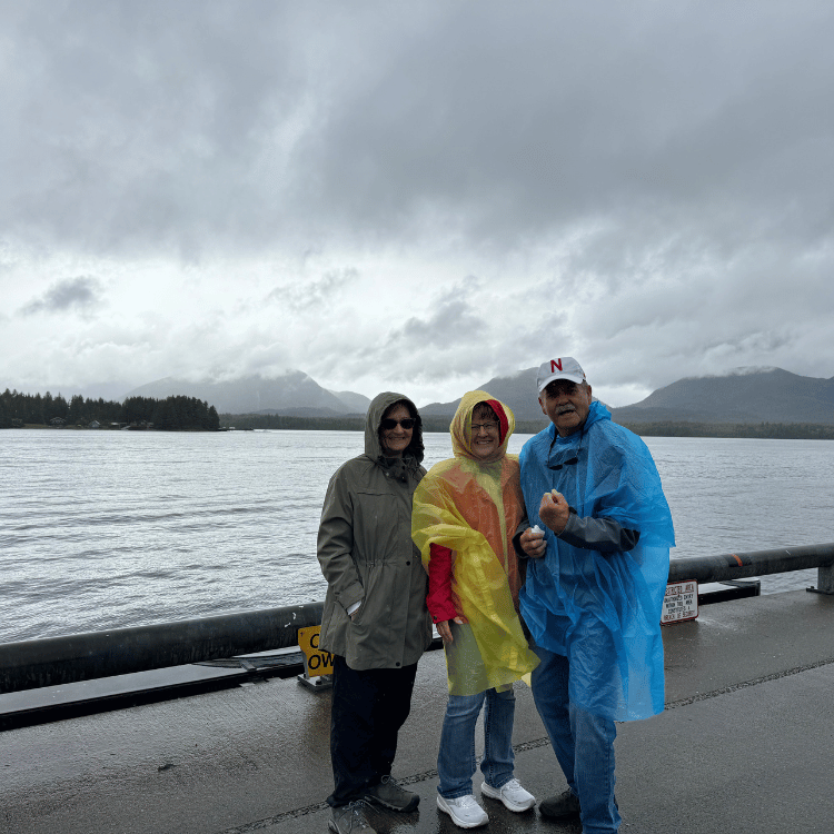 Three people wearing rain gear stand on a dock in front of a body of water under a cloudy sky.