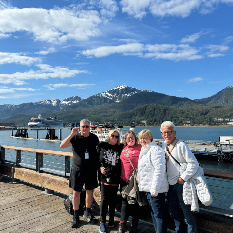Five people pose on a dock in front of a cruise ship and mountainous landscape under a sunny sky.