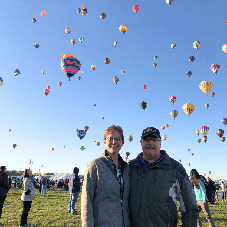Couple smiles at a hot air balloon festival. Many colorful balloons fill the clear blue sky.