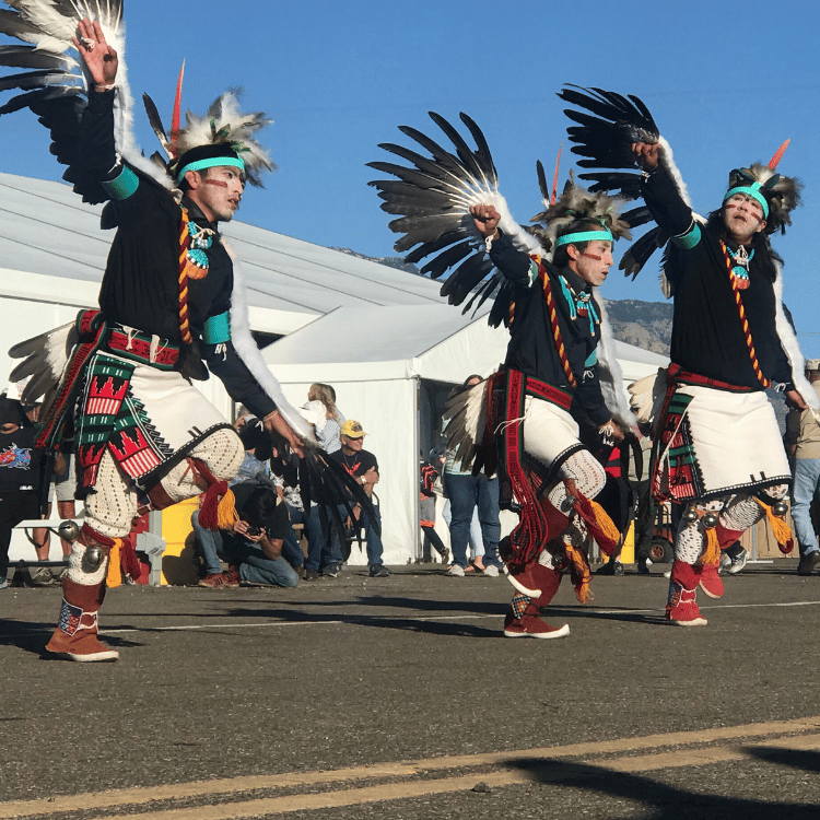 Three Native American dancers in traditional regalia, performing outside under a blue sky.