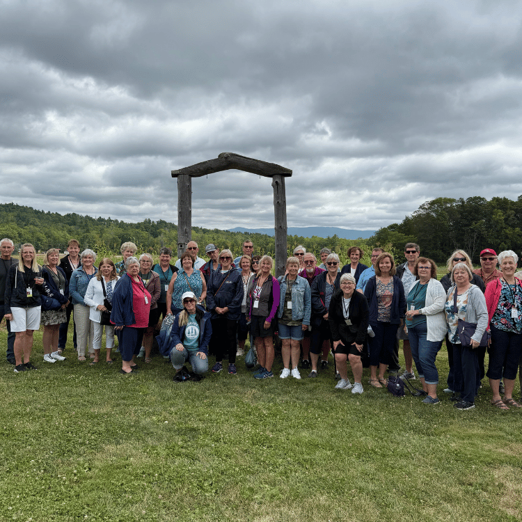 Group of people posing outdoors near a wooden archway, overcast sky, green field, mountains in the background.