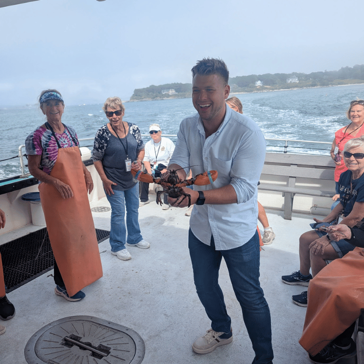 Man on a boat holding lobster, smiling. Women watch in a coastal setting.