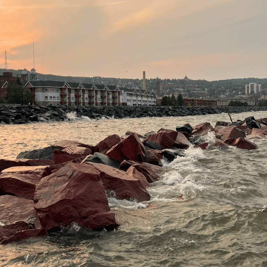 Waves crash over red rocks in a lake, with buildings and a cityscape in the background under an overcast sky.