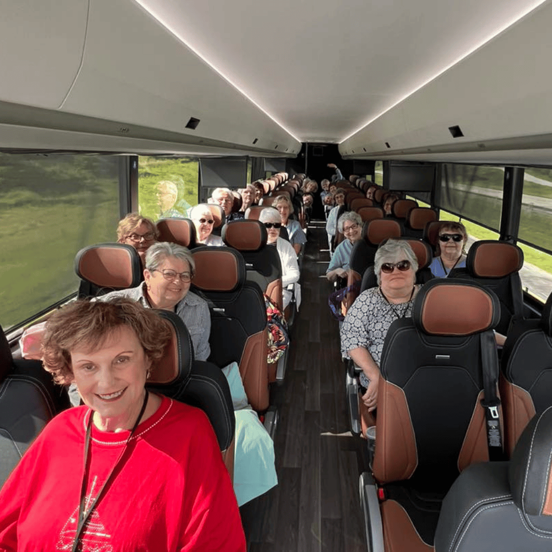 Inside a bus filled with older adults, many wearing sunglasses. Woman in red smiling at camera.