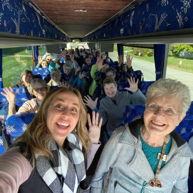 People on a bus, smiling and waving. Two women in the foreground take a selfie, with rows of passengers behind them.