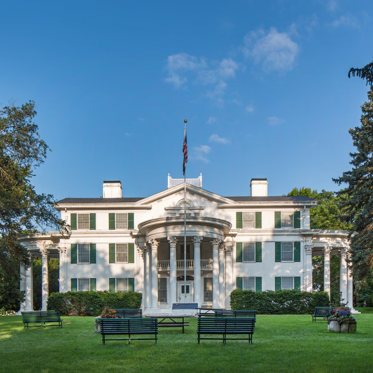 White, two-story mansion with columns, green shutters, flag, benches on lawn, blue sky.